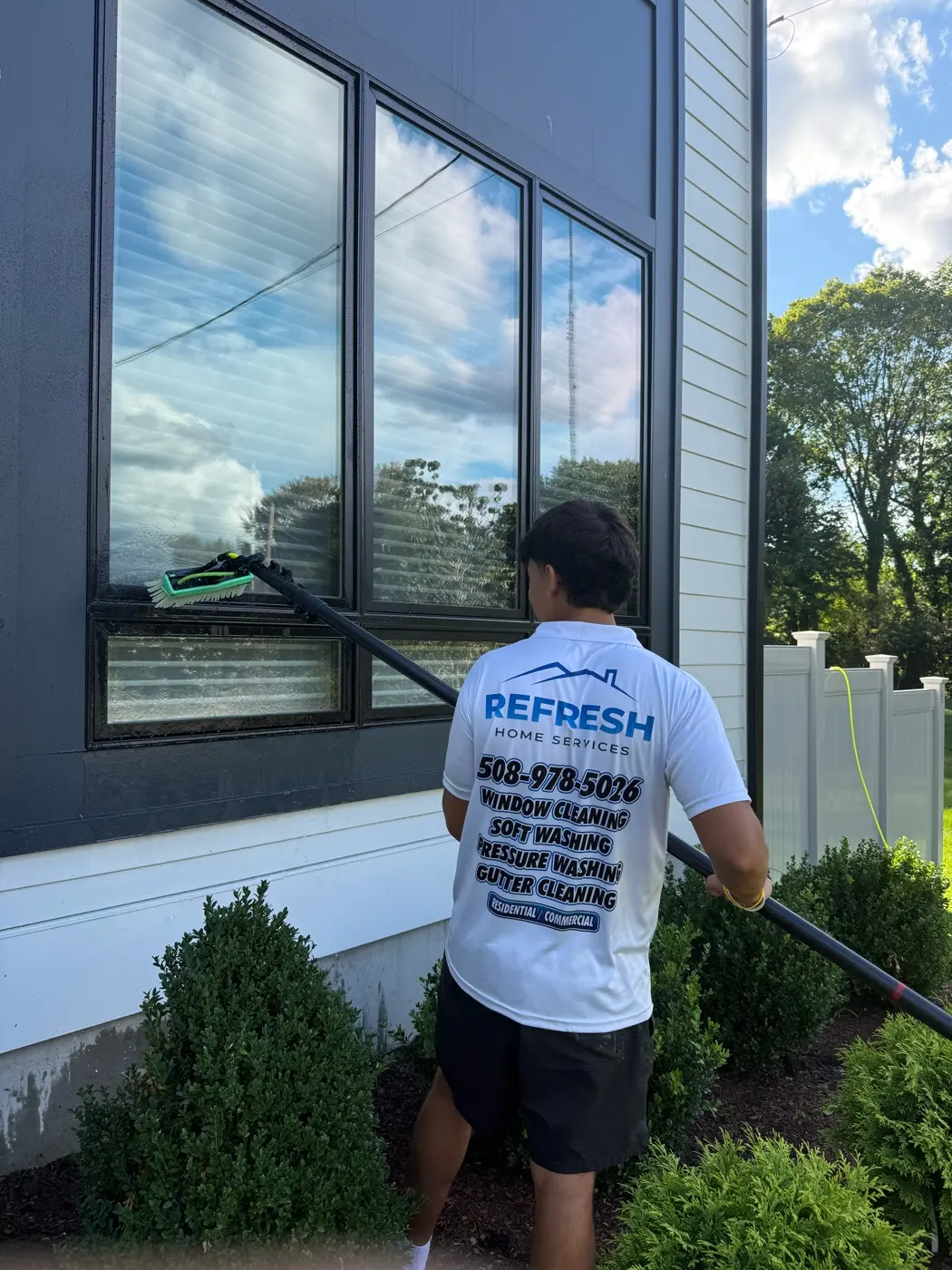 ReFresh Home Services in a branded shirt cleaning a large black-framed window with a water-fed brush pole